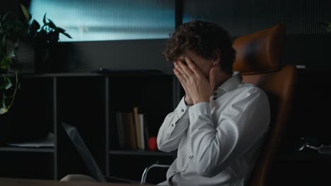 Side-view-of-a-tired-guy-with-curly-hair-in-a-shirt-getting-tired-and-touching-his-face-while-working-on-a-laptop-in-the-office