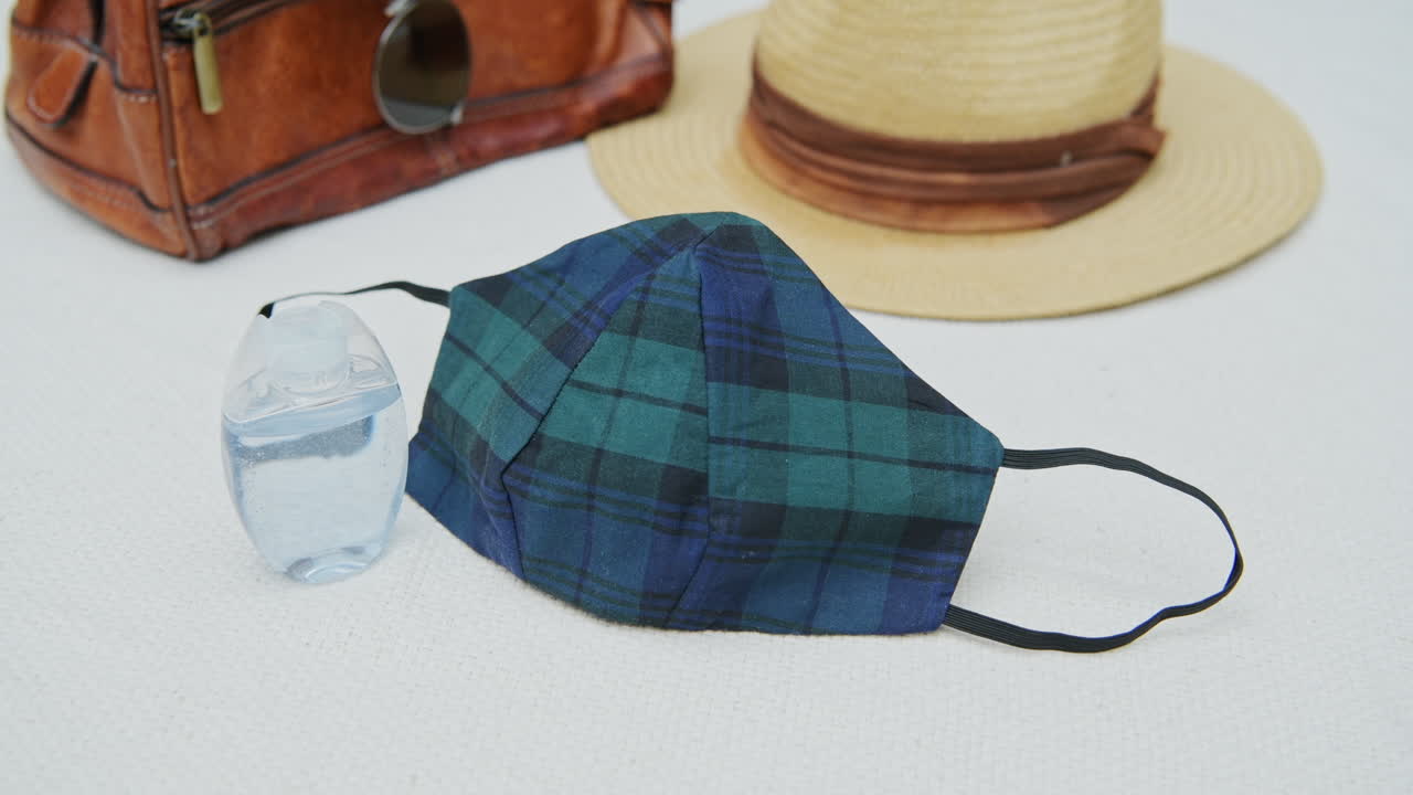 A patterned facemask rotates on a pure white table next to a brown travel bag and sun hat