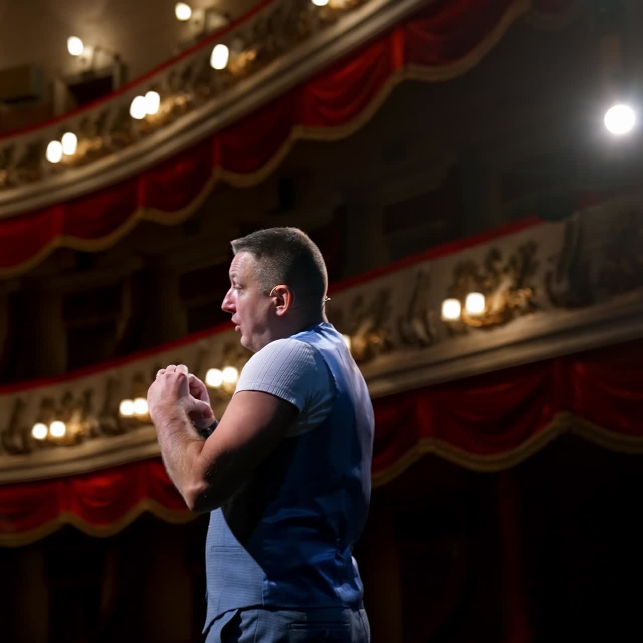 Presenter in front of empty theatre hall with red velvet chairs. Actor is standing on stage and talking with gestures before the performance