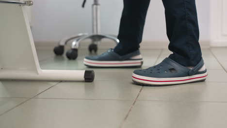 leg view showing skin therapist standing beside massage chair in spa room wearing crocs and white socks on tiled floor next to equipment trolley and lamp in calm professional treatment setting
