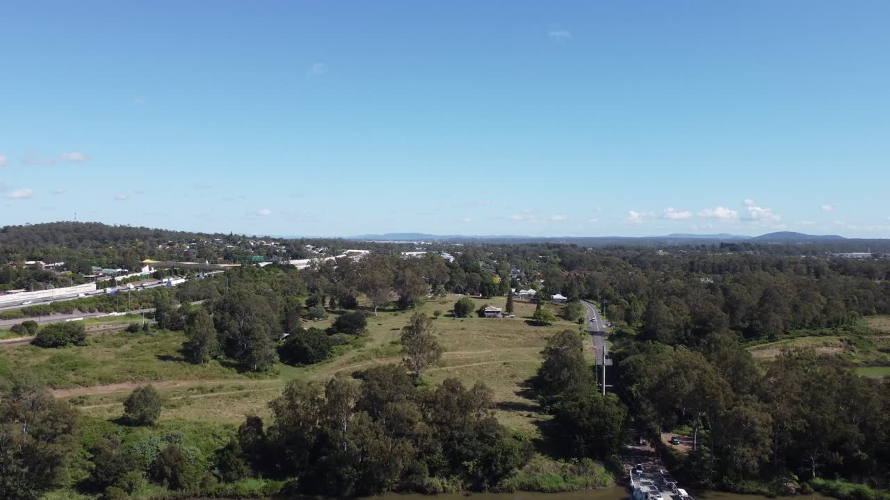 Drone ascending over a brown river showing a ferry and a major highway on the other side of the river
