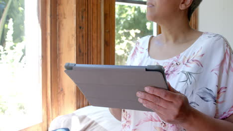 Mujer-Birracial-Mayor-Pensativa-Usando-Tableta-En-La-Ventana-De-Casa,-Cámara-Lenta