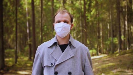 portrait of young guy in protective white medical mask. young guy puts on a face mask to prevent the spread of the virus in city park or forest. pandemic, coronavirus.