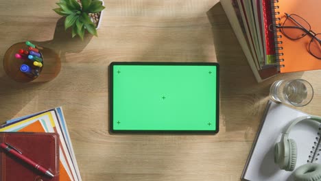 top down view of a laptop computer with mock up green screen chromakey display on a wooden office desk next to notebook with pens, glasses, and a glass of water. slow zoom out, close up
