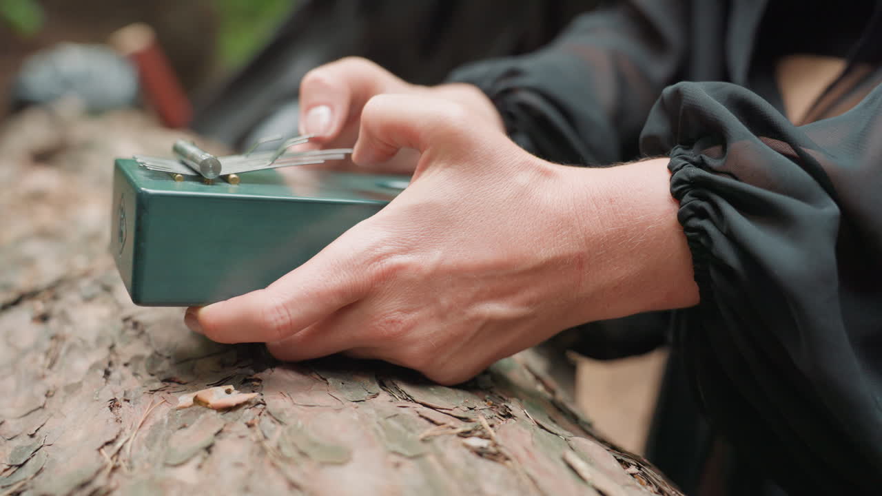 Close up of black fairy hands gently plucking metal tines of green thumb piano resting on rough bark of fallen tree in forest, soft natural light creating magical serene woodland atmosphere