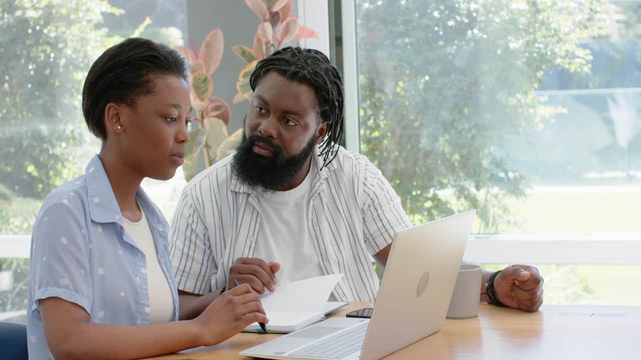 African-American mentor pointing to document, mentee writing on laptop for review by window