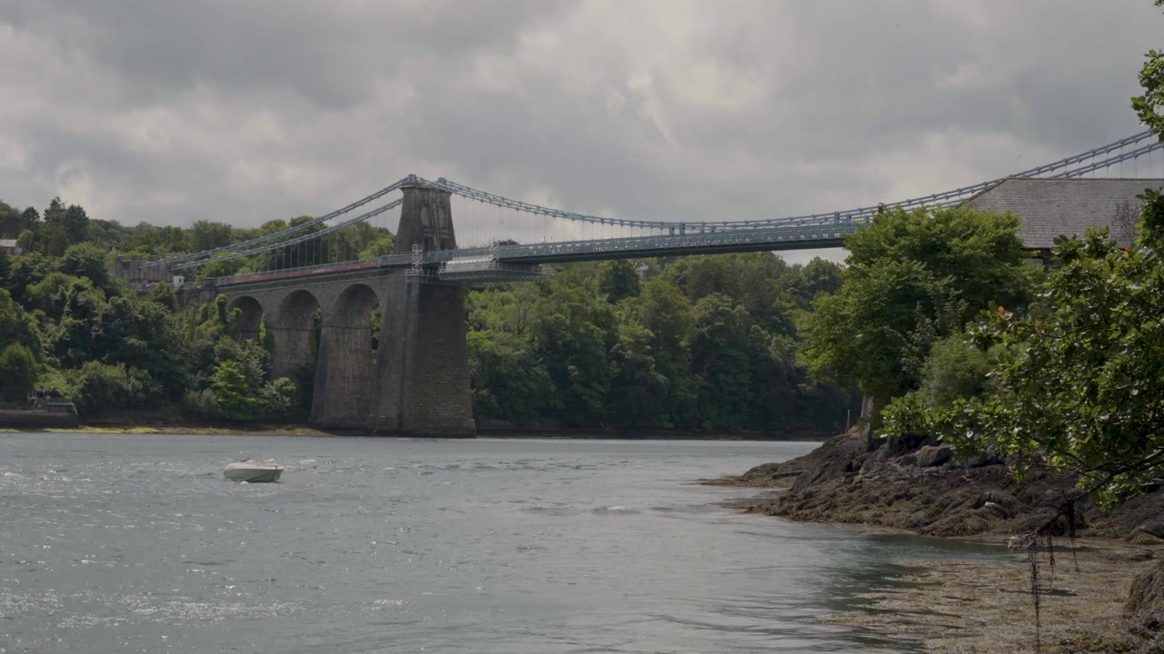 Composizione ad angolo fisso che cattura l'intera lunghezza del Ponte di Menai contro un cielo sereno, enfatizzando la simmetria architettonica e i dettagli della struttura di sospensione.