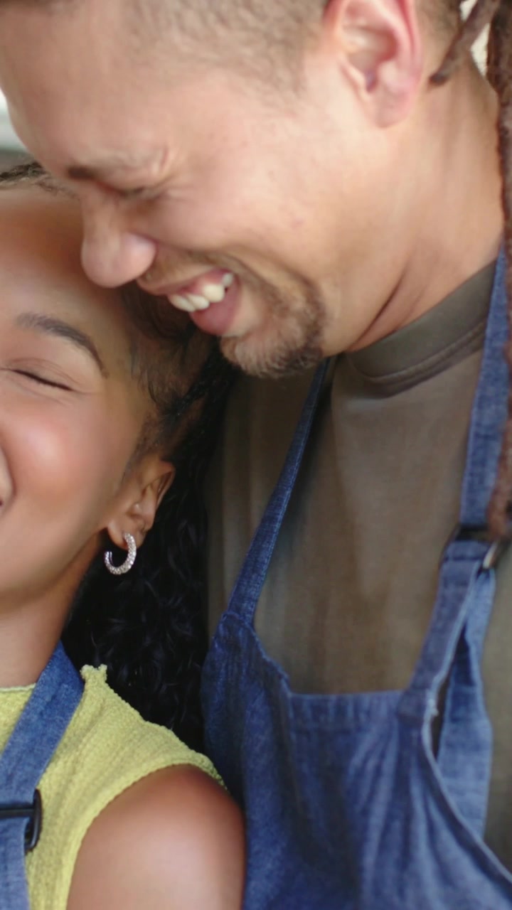 Vertical video: Leaning man nuzzling her temple in kitchen both laughing wearing blue aprons, hoops