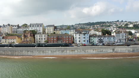 Eine-Profildrohnenaufnahme-Eines-Zuges-Entlang-Der-Dawlish-Stadt-Mit-Häusern-Am-Meer-Im-Hintergrund-Mit-Dem-Meer-Und-Dem-Strand-Im-Schuss