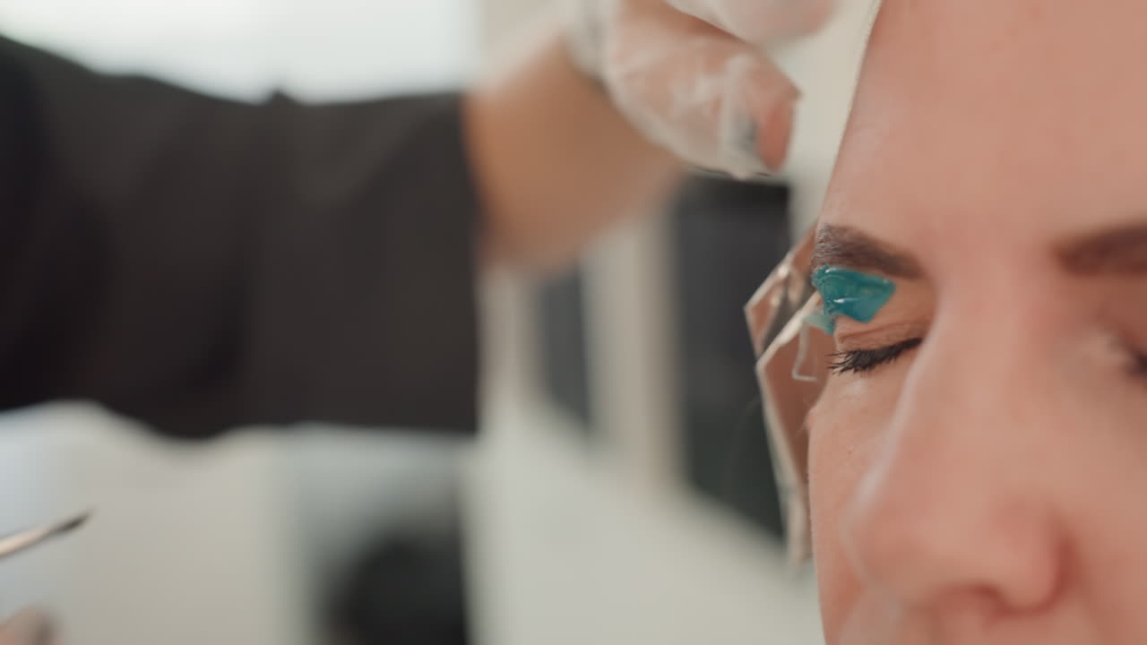 closeup caucasian woman getting teal eyeshadow applied by gloved esthetician in bright salon studio, brush gliding along eyelid, precise pigment placement, relaxed client with closed eyes, soft