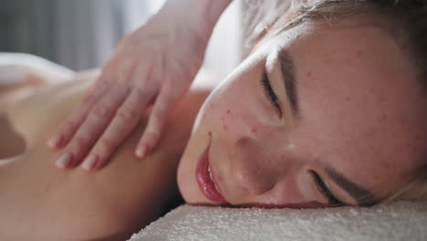 light skin blond woman lying on towel and smiling softly while receiving relaxing shoulder massage from therapist, with close-up focus on her face and hands, illuminated by soft natural light
