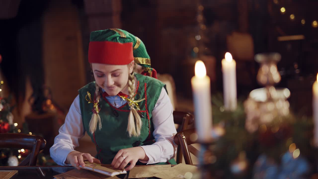 Funny elf with envelopes at the table on festive background. Joyful female elf in green and red costume looking at envelopes for Santa and dances happily.