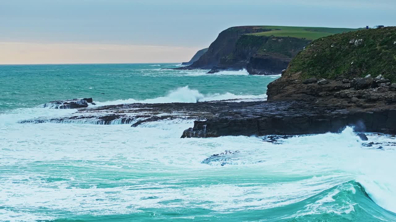 Waves crash and build powerful sprays along rocky shoreline under overcast sky
