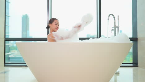 asian woman foaming her body, she is lying in a bathtub full of foam on a high floor of the luxury apartment complex with glass walls in the background