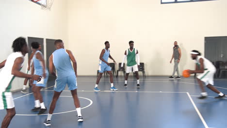 multiracial male basketball players playing basketball, passing with coach observing in gym