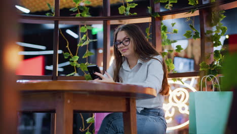 woman operating her phone in a vibrant bar, glancing at something before refocusing on her phone while other shoppers move around in the background, decorated with unique lighting and designs