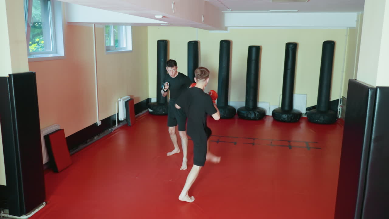 레슬링 선수들이 훈련 체육관 안 빨간색 매트 위에서 스파링을 하고 있어요. 한 선수가 상대를 향해 발차기를 하고 다른 선수는 방어할 준비를 하고 있으며, 둘 다 검은색 운동복과 장갑을 착용하고 집중하고 있어요. 주위에는 검은색 펀칭백이 놓여 있어요.