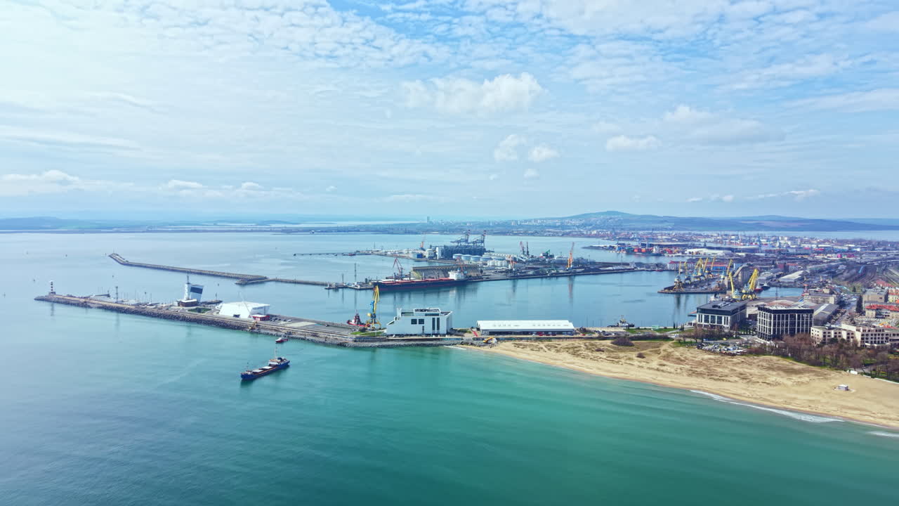 Ombak menghantam pantai di pelabuhan yang sibuk dekat kota di bawah langit berawan pada siang hari dengan kapal dan bangunan terlihat di kejauhan.