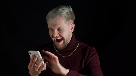 Smiling-shocked-happy-winning-of-man-with-blue-eyes-while-browsing-on-smartphone-on-black-background