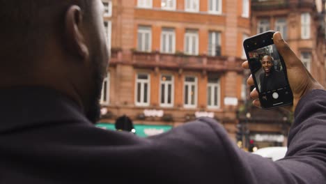 Male-taking-a-selfie-photo-and-smiling-at-his-smart-phone-on-a-street-in-London-City