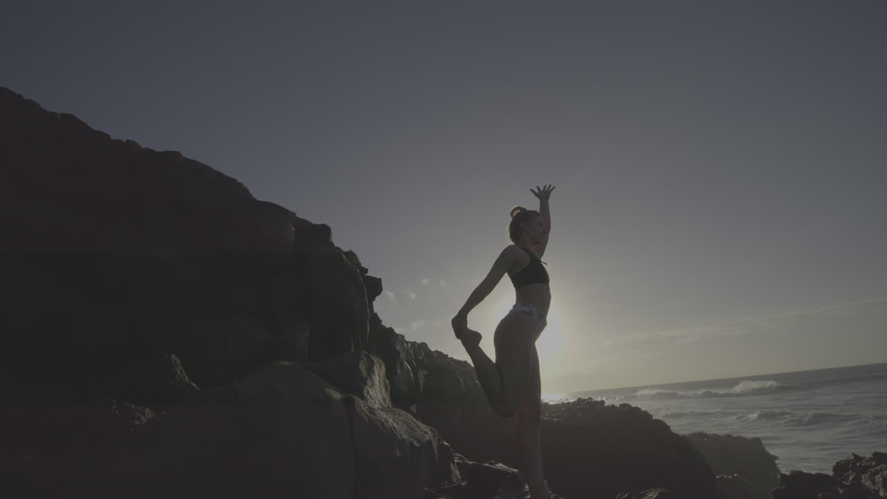 Woman practicing yoga on a rocky beach at sunset