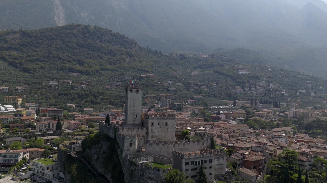 Aerial drone shot flying from Lake Garda toward the historic town of Malcesine, focusing on the iconic Scaliger Castle. Beautiful lakeside scenery with mountains and clear blue water.