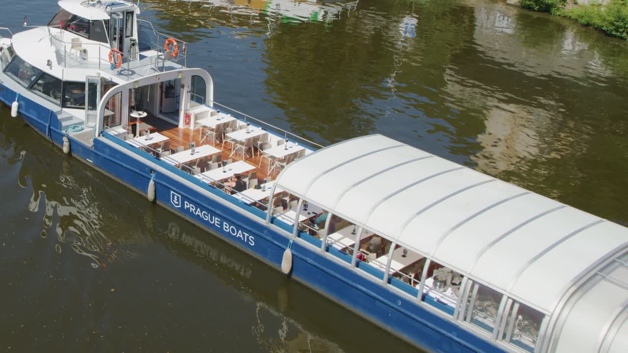 A modern sightseeing boat with a retractable roof cruises along the Vltava River in Prague on a sunny day, filmed from above with smooth camera movement