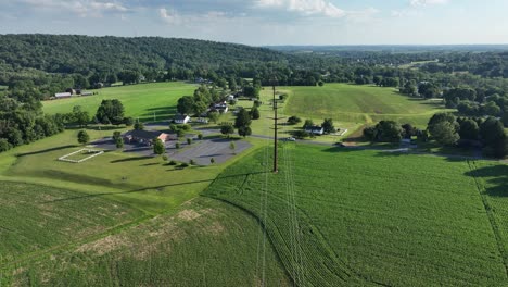 Fliegen-über-Die-üppig-Grüne-Ländliche-Landschaft-Mit-Feldern-Und-Kleinen-Häusergruppen-An-Einem-Sonnigen-Tag-Mit-Einer-Stromleitung-In-Der-Mitte