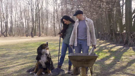 cucasian couple and their dog holding a wheelbarrow and a rake in the countryside