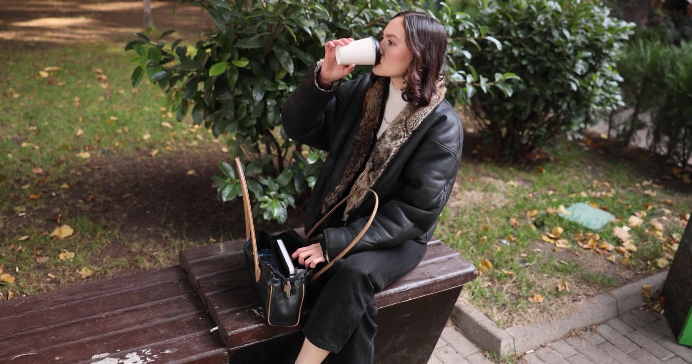 Donna beve caffè cercando il telefono nella borsa seduta su una panchina
