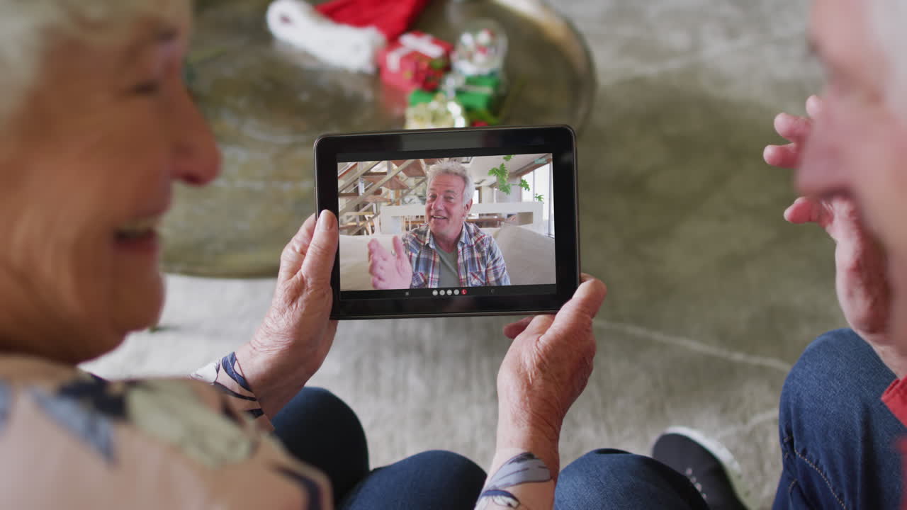 Coppia anziana caucasica sorridente che usa un tablet per una videochiamata di Natale con un uomo sullo schermo.