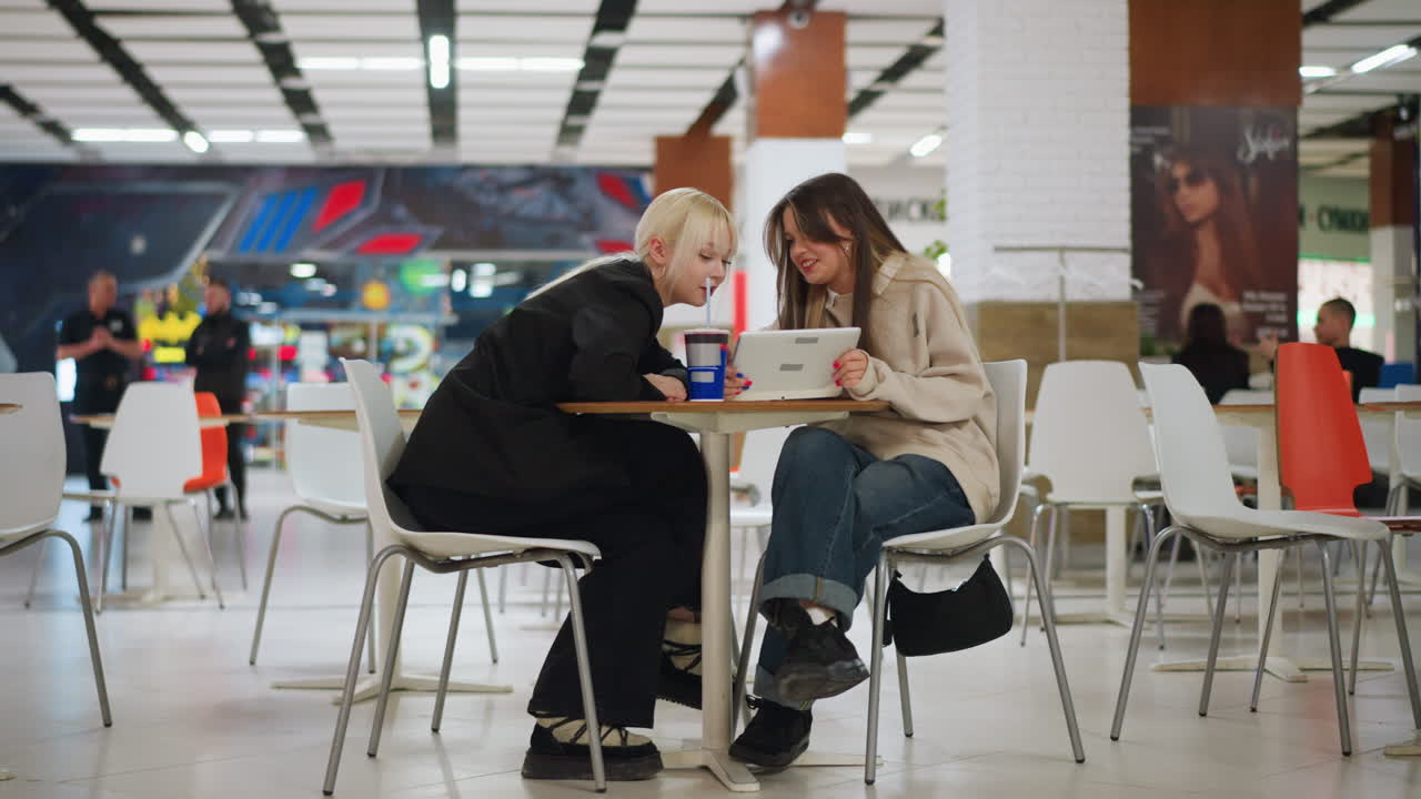 Due giovani donne sedute a un tavolo in un bar luminoso di un centro commerciale, una tiene in mano un tablet mentre l'amica vestita di nero si sporge per guardare, entrambe condividono interazione e comunicazione in un contesto di stile di vita moderno