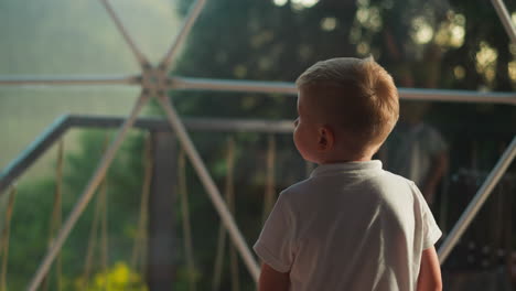 little child enjoys sunset in wild nature looking out window. toddler dreams looking at landscape calmly on blurred background backside view slow motion