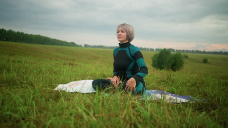 middle-aged woman seated on mat in an accomplished pose, practicing yoga in vast grassy field with overcast sky, surrounded by nature, she holds her position in a serene, focused posture