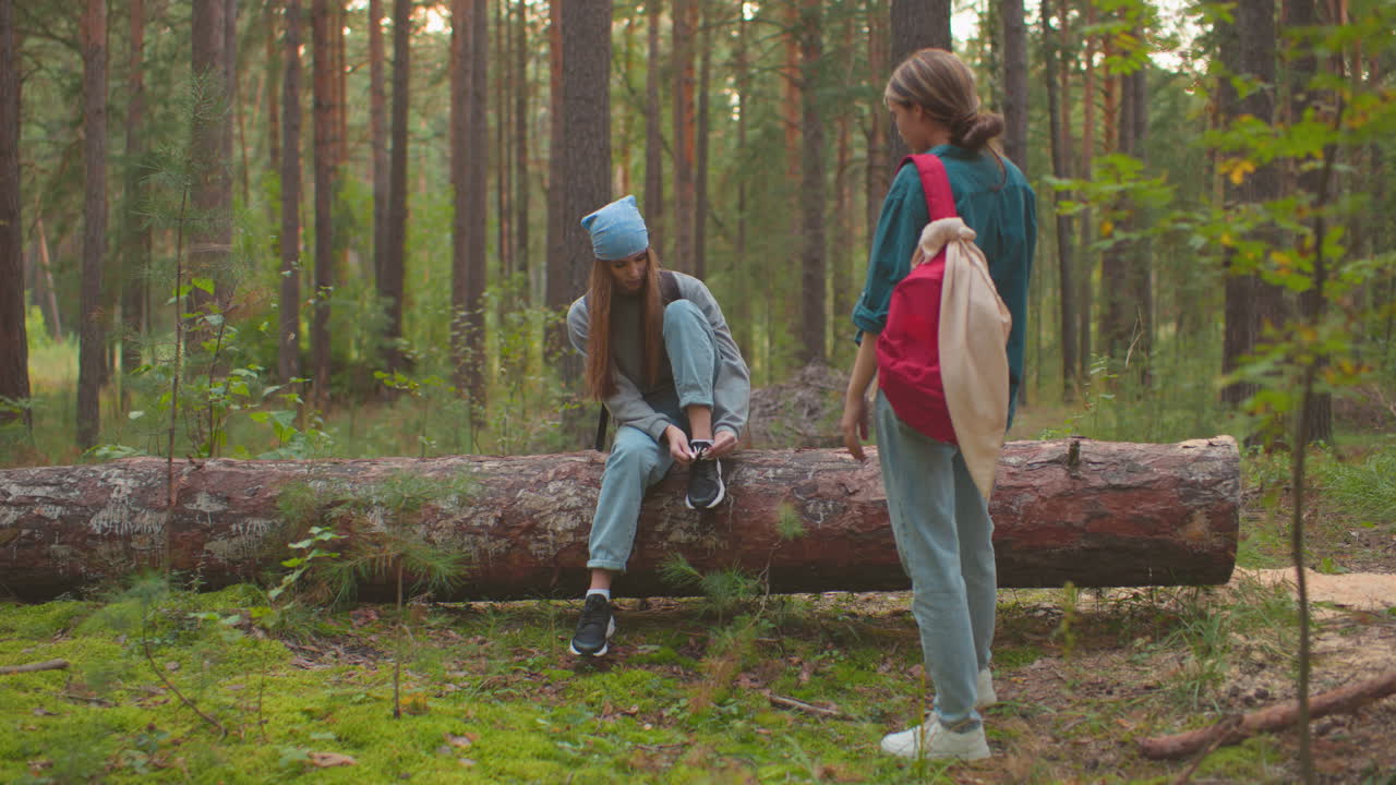 Siblings take a break in serene forest surroundings, one sitting on a fallen tree, tying her shoe with a blue bandana on her head, while her companion stands close, engaging in conversation