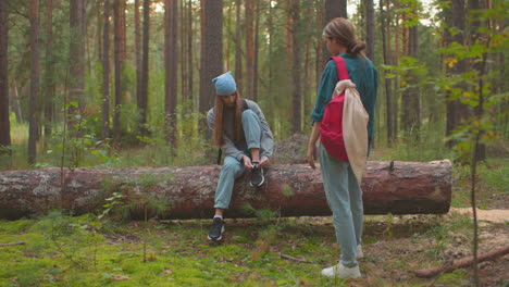 siblings take a break in serene forest surroundings, one sitting on a fallen tree, tying her shoe with a blue bandana on her head, while her companion stands close, engaging in conversation