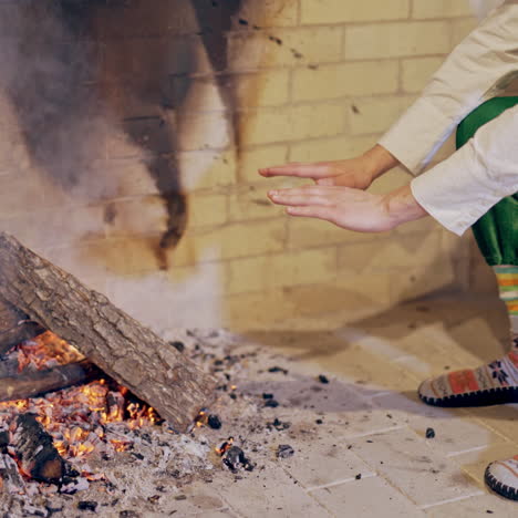 elf is sitting near the fire and warming his cold hands in the house. young man elf rubbing his cold hands near the fireplace. fairy character elf is heating hands on christmas.