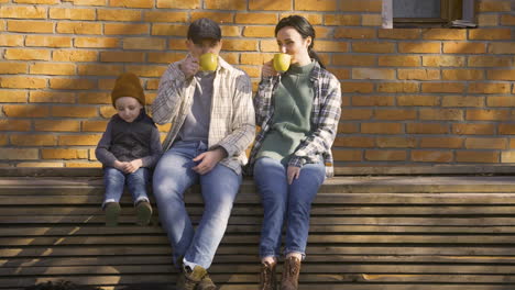 Caucasian-couple-talking-with-her-son-while-they-drinking-tea-outside-a-country-house.-Their-dog-walks-around-them