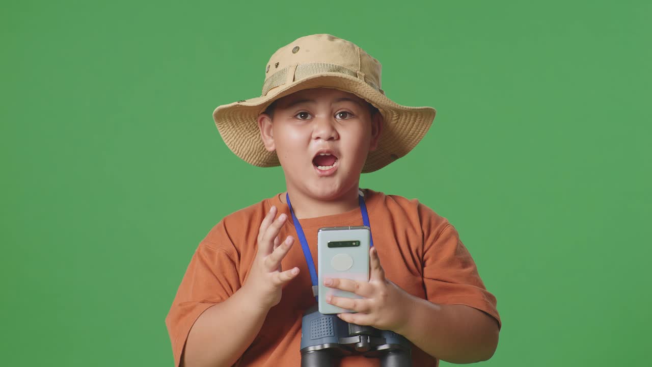 모자와 안경을 쓴 아시아 관광 소년은 스마트폰을 보고 와우라고 말합니다. 소년 연구자는 녹색 화면 배경에서 무언가를 조사합니다. 여행, 관광, 모험 개념, 근접