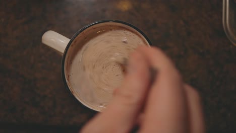 top view of creamy hot chocolate drink in a mug stirred with a teaspoon in the kitchen - overhead shot