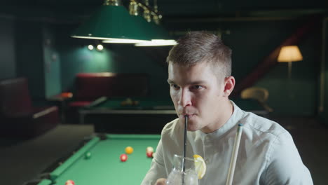 close-up of billiard player wearing white shirt, sitting on green pool table, holding cue stick and glass of lemon drink with black straw. he raises the glass and takes a sip, with focused expression