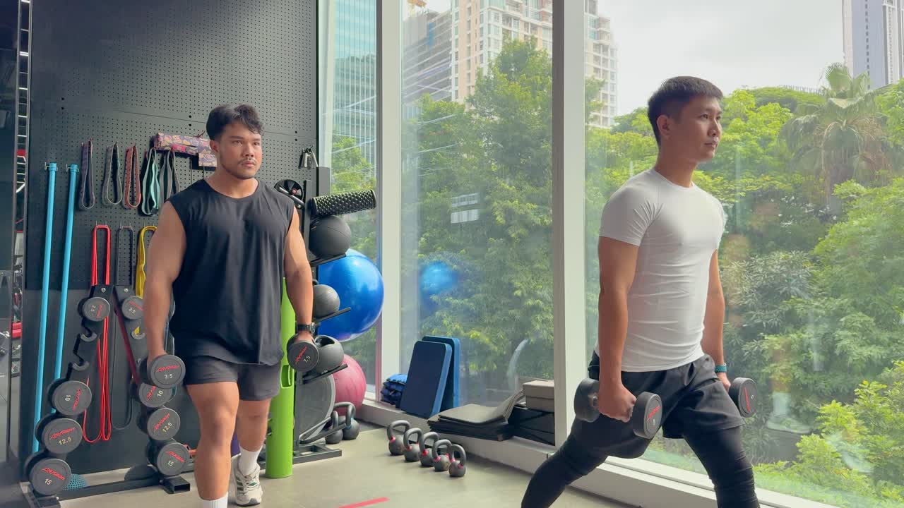 Two men perform lunges with dumbbells in a gym, surrounded by fitness equipment, against a backdrop of large windows