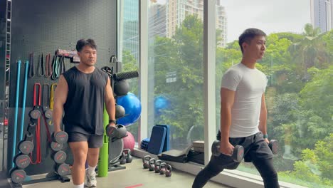 two men perform lunges with dumbbells in a gym, surrounded by fitness equipment, against a backdrop of large windows