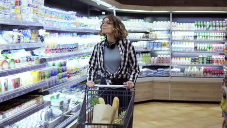 Modern-young-carefree-girl-in-glasses-and-headphones-on-neck-walking-with-shopping-cart-in-the-supermarket-and-steering-around