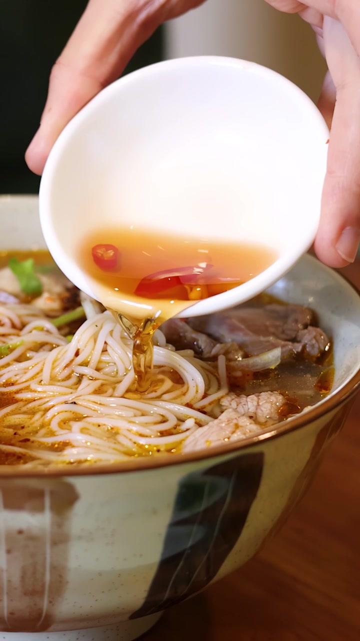 Primo piano di mani che versano salsa piccante in una ciotola di ceramica di zuppa di noodle tradizionale
