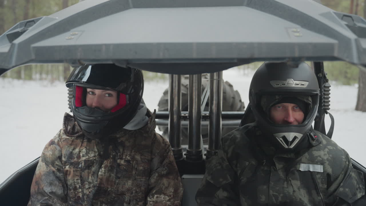 Due piloti bianchi, con caschi e giacche mimetiche, siedono nella cabina di un ATV/UTV dietro il parabrezza in una foresta di pini innevata. I motori sono al minimo, il respiro è visibile. C'è tensione e cameratismo mentre si preparano per un'impresa estrema.