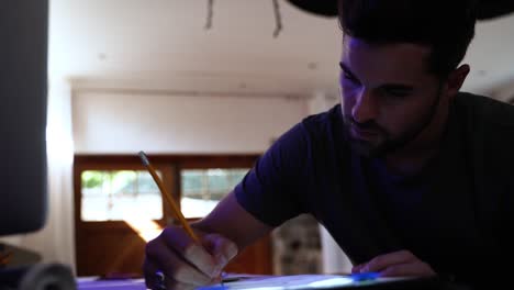 joven hombre guapo con cabello oscuro y barba sentado en su escritorio dibujando en papel con un lápiz y mostrando que está pensando trayendo su mano a su frente por un momento