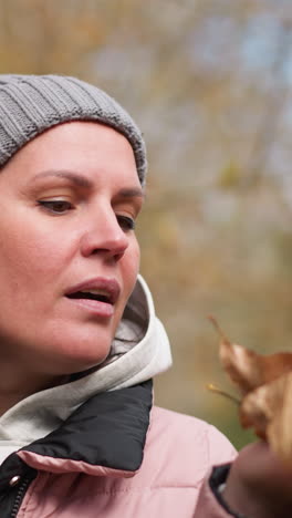 mujer con gorra gris y chaqueta rosa soplando follaje seco en su mano, rodeada de árboles de otoño borrosos en el fondo, creando una atmósfera de otoño pacífica