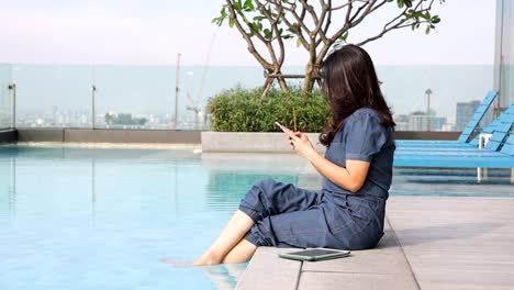 asian woman using smartphone share photos, communicate with friends on vacation, sitting on the edge dangling her feet in swimming pool on skyscraper, woman looking at cityscape while relaxing.