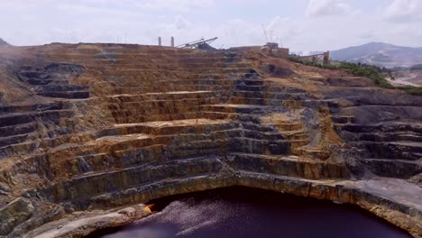 open-pit gold and silver mine, colorful mineral-stained rock walls and a dark, contaminated tailings lake, barrick gold, cotuí, dominican republic. aerial forward